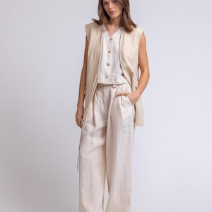 May include: A three-piece linen outfit in a neutral beige color. The ensemble includes wide-leg pants, a button-down top, and a sleeveless vest. The model is wearing white sandals. The outfit is made of a lightweight fabric.