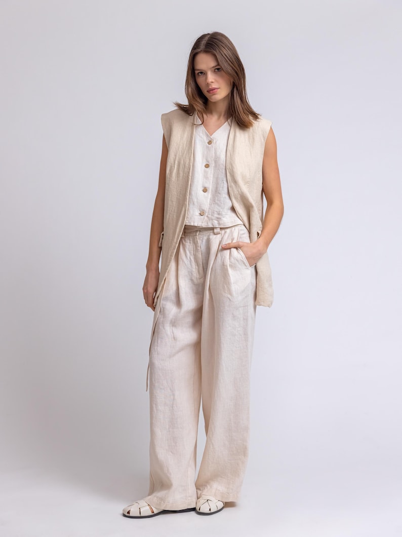 May include: A three-piece linen outfit in a neutral beige color. The ensemble includes wide-leg pants, a button-down top, and a sleeveless vest. The model is wearing white sandals. The outfit is made of a lightweight fabric.