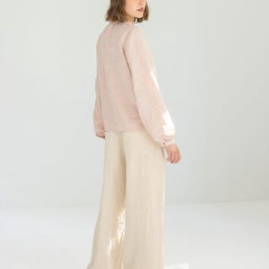 May include: A light pink long-sleeve blouse with a round neckline and buttoned cuffs. The blouse is paired with wide-leg, cream-colored pants. The outfit is made of a lightweight fabric, suitable for warmer weather. The model is wearing black shoes.