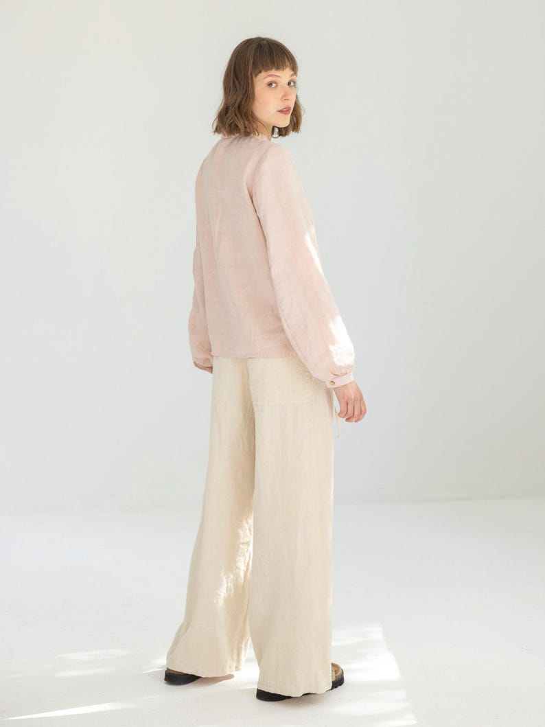May include: A light pink long-sleeve blouse with a round neckline and buttoned cuffs. The blouse is paired with wide-leg, cream-colored pants. The outfit is made of a lightweight fabric, suitable for warmer weather. The model is wearing black shoes.