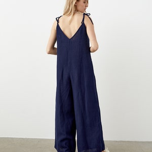 May include: A navy blue jumpsuit with a V-neck and wide legs. The straps are tied at the shoulders. The model is wearing white sneakers. The jumpsuit is made of a lightweight fabric.
