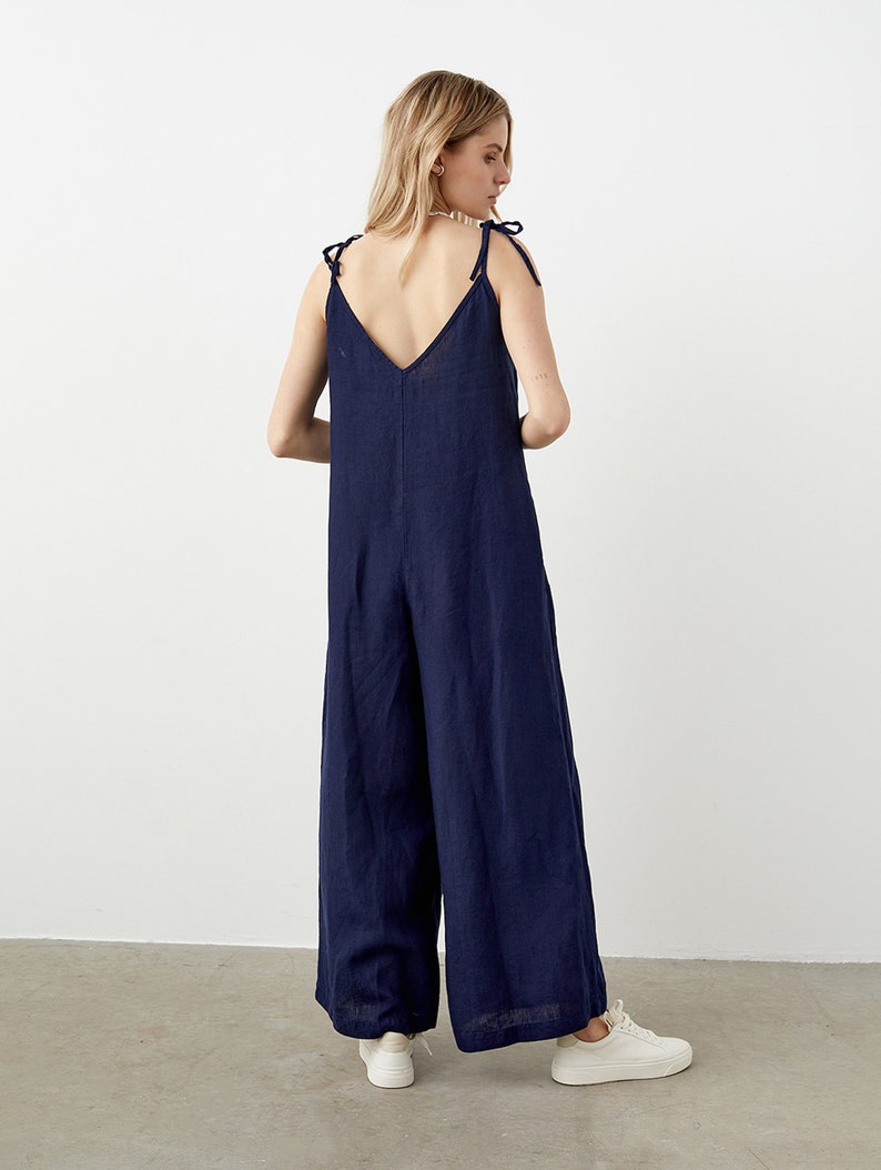 May include: A navy blue jumpsuit with a V-neck and wide legs. The straps are tied at the shoulders. The model is wearing white sneakers. The jumpsuit is made of a lightweight fabric.
