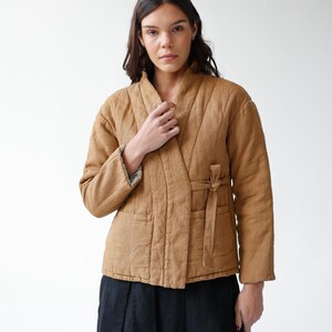 May include: A tan quilted jacket with a wrap-around design and a tie closure. The jacket has a shawl collar and two front pockets. The model is wearing a black skirt. The jacket is made of a soft, textured fabric.