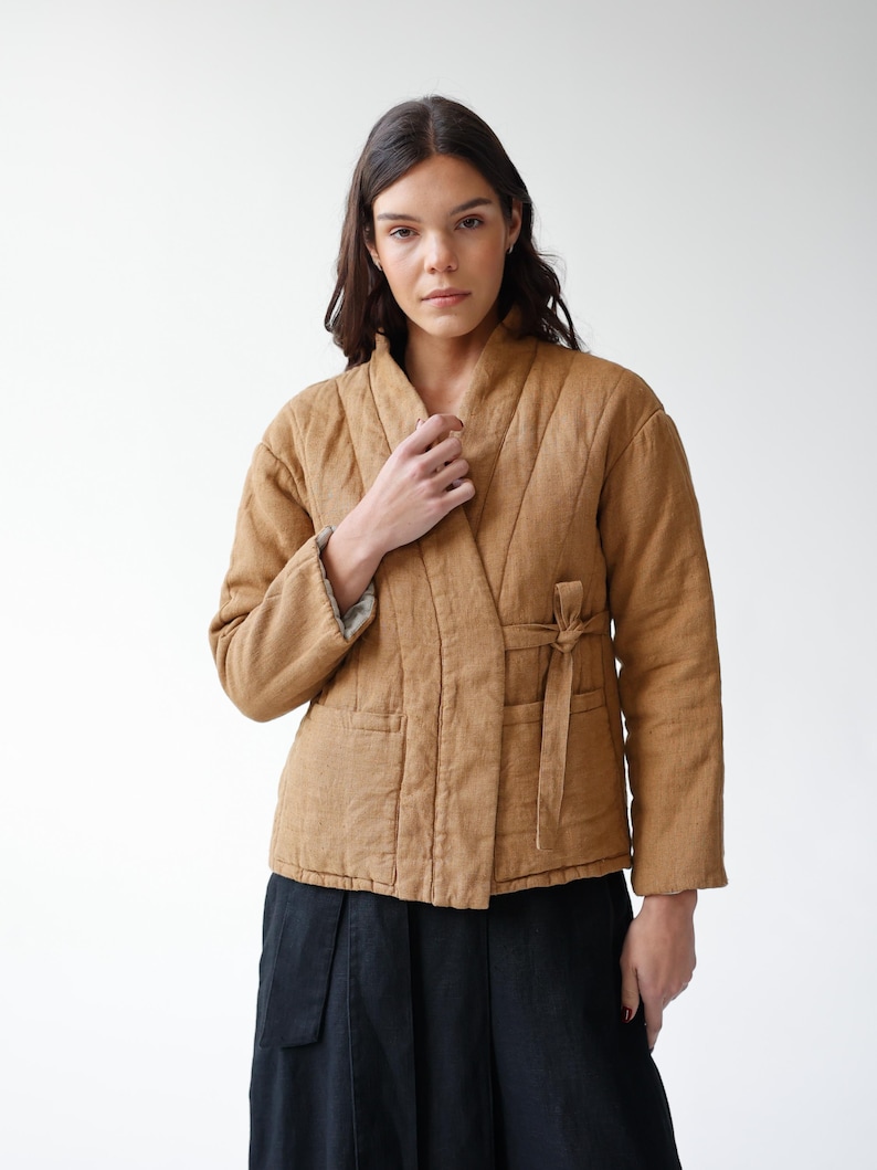 May include: A tan quilted jacket with a wrap-around design and a tie closure. The jacket has a shawl collar and two front pockets. The model is wearing a black skirt. The jacket is made of a soft, textured fabric.
