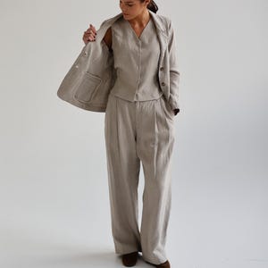 May include: A three-piece linen suit in a neutral beige color. The suit includes a blazer, a vest, and wide-leg trousers. The blazer has two buttons and a pocket. The vest has a V-neck. The trousers are pleated.