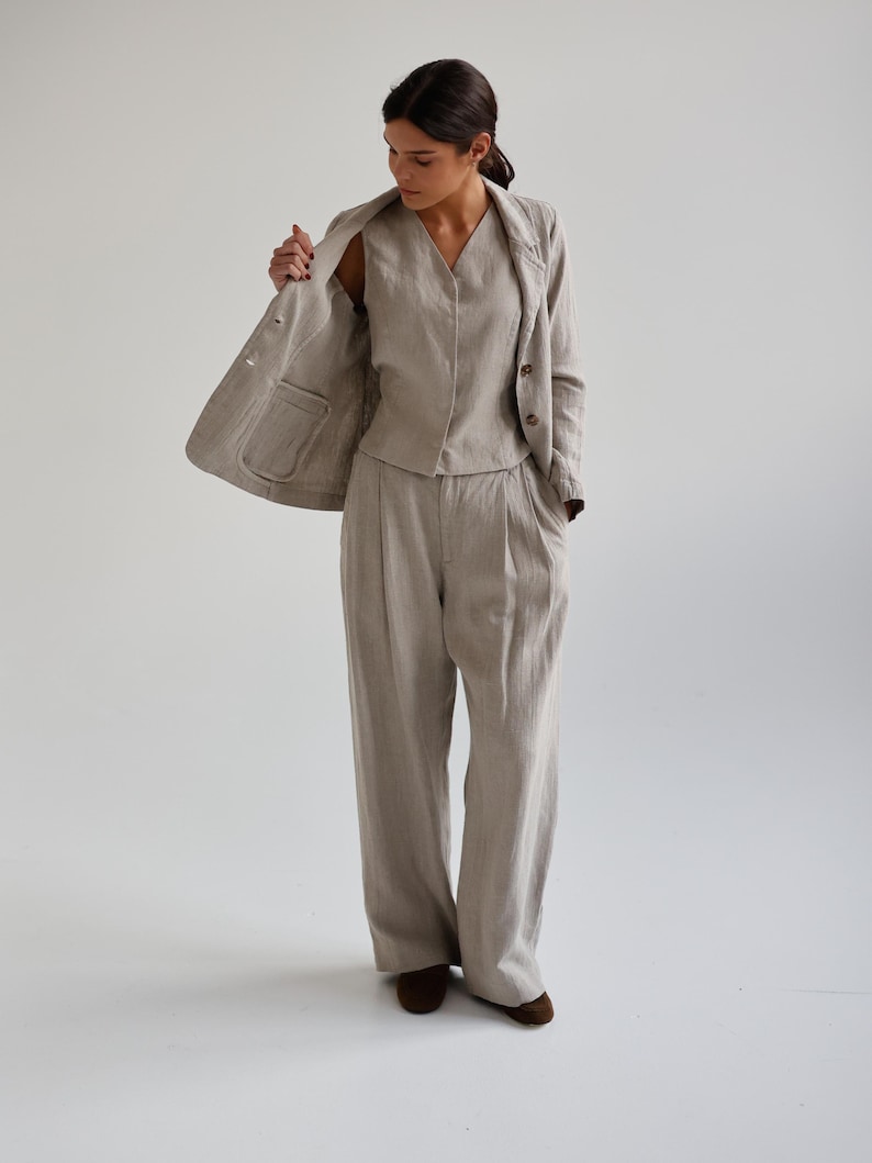 May include: A three-piece linen suit in a neutral beige color. The suit includes a blazer, a vest, and wide-leg trousers. The blazer has two buttons and a pocket. The vest has a V-neck. The trousers are pleated.