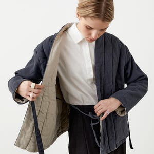 May include: A navy blue quilted jacket with a light beige lining. The jacket has a tie closure and is worn over a white button-down shirt and black pants. The jacket's design features vertical stitching and a relaxed fit.