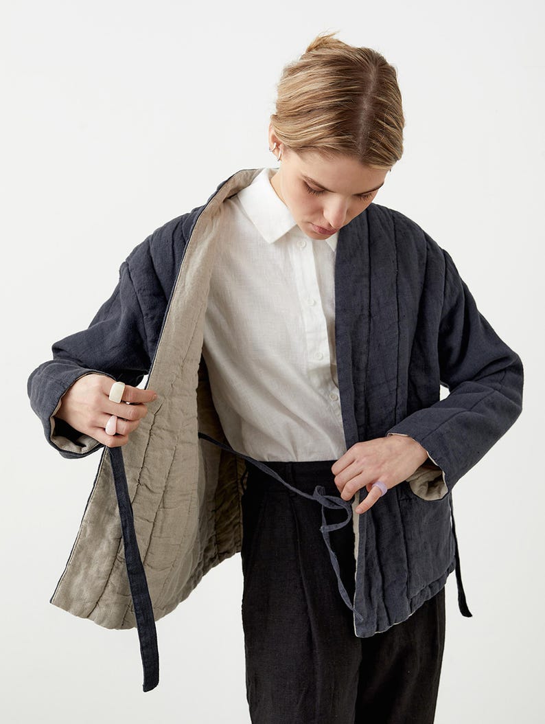 May include: A navy blue quilted jacket with a light beige lining. The jacket has a tie closure and is worn over a white button-down shirt and black pants. The jacket's design features vertical stitching and a relaxed fit.