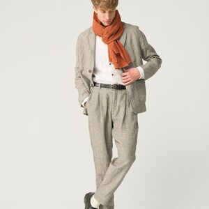 May include: A light gray linen suit with a matching jacket and pleated pants. The outfit includes a white button-down shirt, a rust-colored scarf, and a black leather belt. Black leather shoes complete the look.