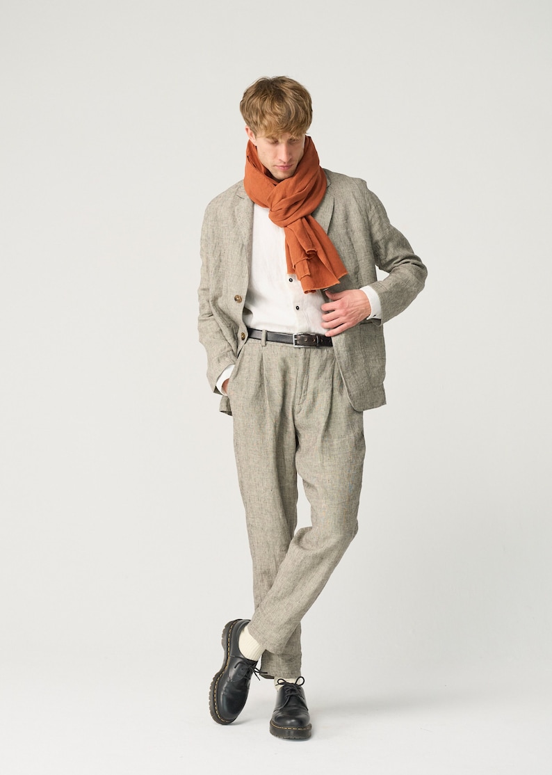 May include: A light gray linen suit with a matching jacket and pleated pants. The outfit includes a white button-down shirt, a rust-colored scarf, and a black leather belt. Black leather shoes complete the look.
