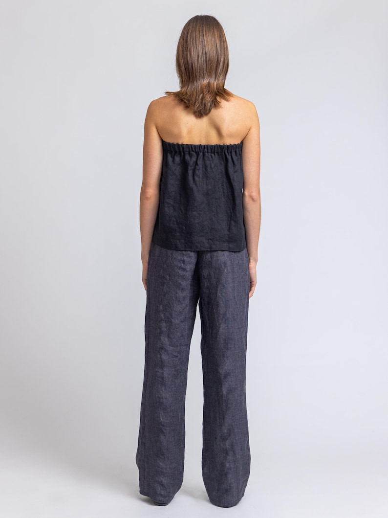 May include: Black strapless top and wide-leg pants. The top has a gathered neckline. The pants are a dark gray color. The outfit is made of a lightweight fabric, perfect for warm weather. The model is facing away from the camera.
