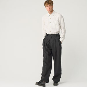 May include: A man wearing a white striped long-sleeve shirt and black pleated pants. He is standing with his hands in his pockets and looking at the camera.