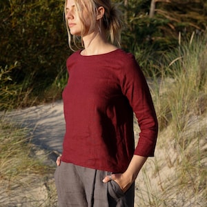 Può includere: Una camicia bordeaux a maniche lunghe e pantaloni grigi. La camicia ha uno scollo tondo e maniche a tre quarti. I pantaloni sono ampi con tasche. L'outfit è realizzato in un tessuto morbido e naturale.