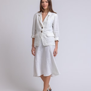 May include: A white linen blazer with rolled-up sleeves and a matching knee-length skirt. The blazer has two front pockets and brown buttons. The outfit is paired with white sandals with black soles. The ensemble is a classic, elegant look.