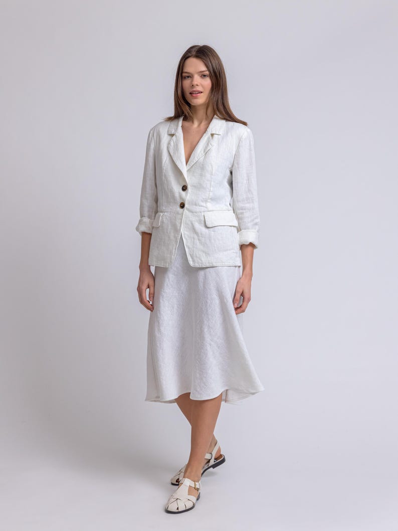 May include: A white linen blazer with rolled-up sleeves and a matching knee-length skirt. The blazer has two front pockets and brown buttons. The outfit is paired with white sandals with black soles. The ensemble is a classic, elegant look.