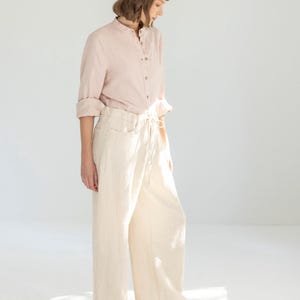 May include: A light pink linen shirt with a mandarin collar and button closure, paired with cream-colored wide-leg pants. The pants have a drawstring waist and pockets. Beige sneakers complete the outfit, creating a casual, comfortable look.