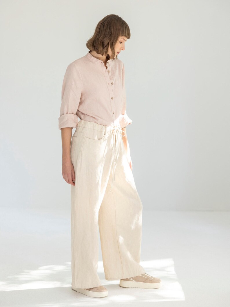 May include: A light pink linen shirt with a mandarin collar and button closure, paired with cream-colored wide-leg pants. The pants have a drawstring waist and pockets. Beige sneakers complete the outfit, creating a casual, comfortable look.