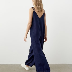 May include: A navy blue jumpsuit with a V-neck and wide legs. The jumpsuit has tie straps and is paired with white sneakers. The model is walking away from the camera, showcasing the back of the garment.