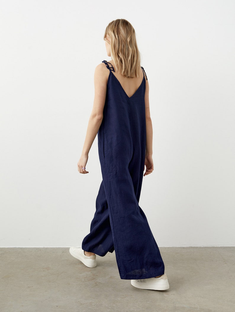 May include: A navy blue jumpsuit with a V-neck and wide legs. The jumpsuit has tie straps and is paired with white sneakers. The model is walking away from the camera, showcasing the back of the garment.