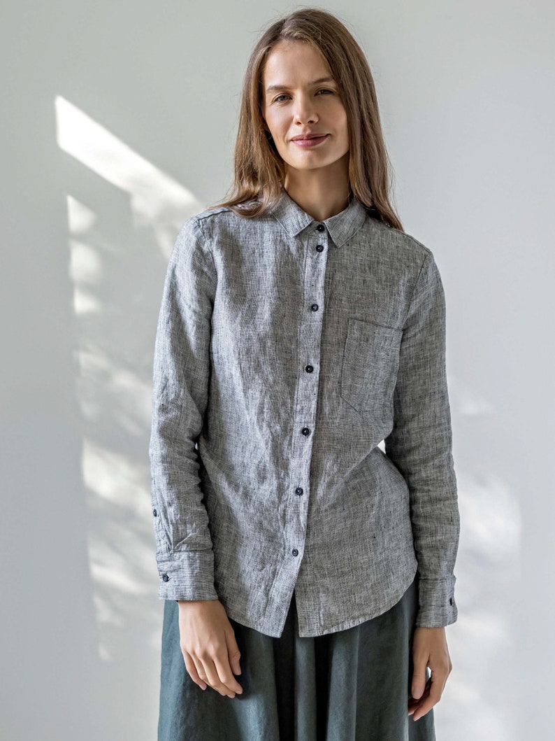 May include: A gray linen button-down shirt with long sleeves and a chest pocket. The shirt has black buttons and is worn with a dark green skirt. The fabric has a textured appearance.