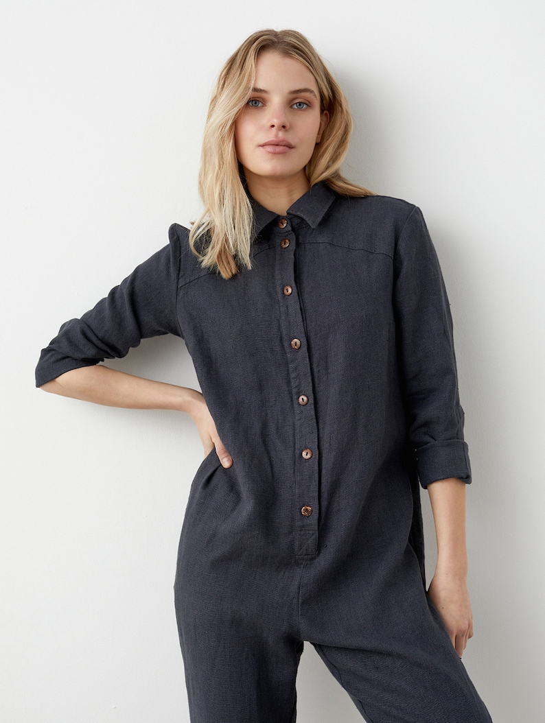 May include: A dark gray linen jumpsuit with a button-up front and long sleeves. The jumpsuit has a relaxed fit and a slightly cropped leg.