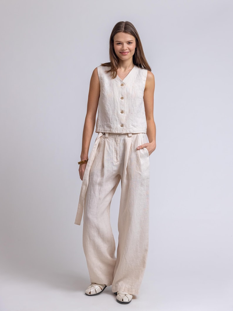 May include: A beige linen two-piece outfit. The sleeveless top has a V-neck and wooden buttons. The high-waisted, wide-leg pants have a matching fabric belt. The outfit is made of a lightweight, textured fabric, perfect for warm weather.