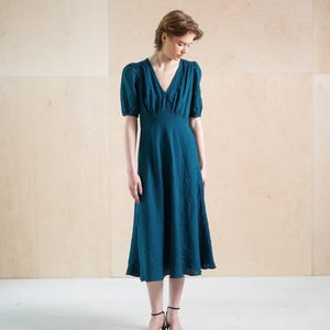 Może przedstawiać: Tealowa sukienka midi z dekoltem w serek i krótkimi, bufiaste rękawami. Sukienka ma dopasowany gorset i zwiewną spódnicę. Modelka ma na sobie czarne sandały na obcasie. Tło to jasny panel drewniany.