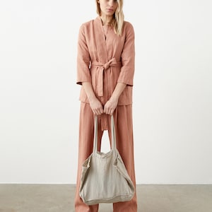 May include: A woman wearing a salmon-colored linen outfit, including a belted jacket and wide-leg pants. She holds a large, natural-colored linen tote bag. The outfit is paired with brown shoes. The background is a plain white wall.