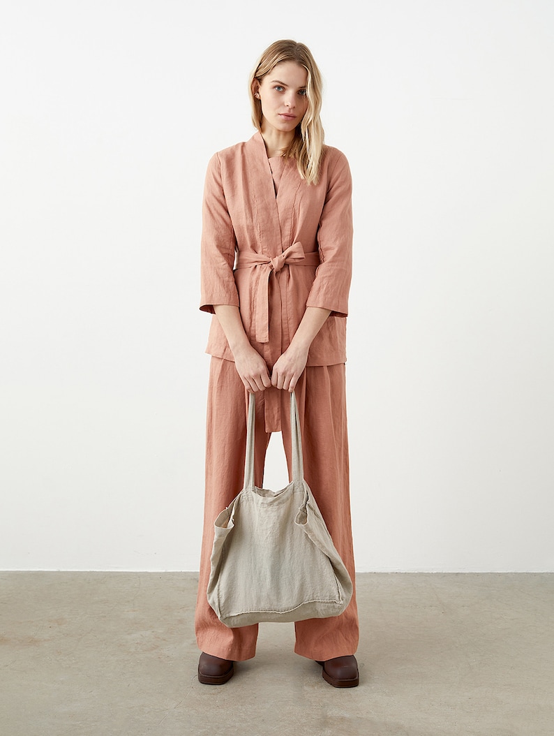 May include: A woman wearing a salmon-colored linen outfit, including a belted jacket and wide-leg pants. She holds a large, natural-colored linen tote bag. The outfit is paired with brown shoes. The background is a plain white wall.