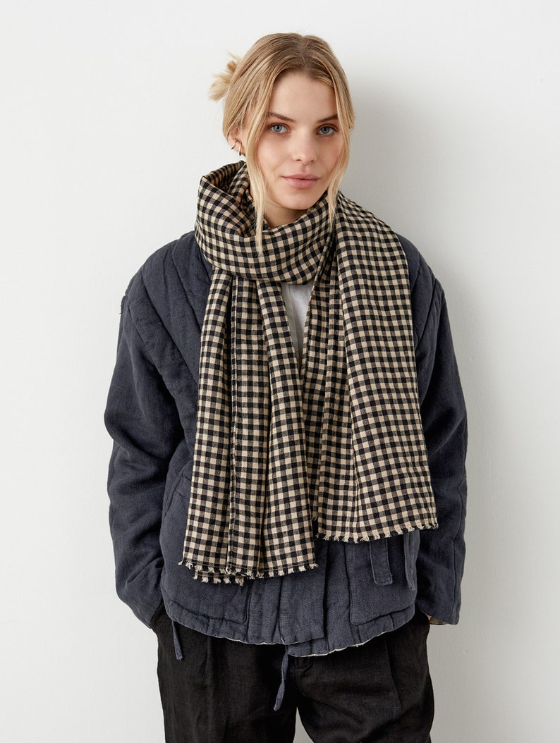 May include: A black and beige gingham scarf with frayed edges. The scarf is worn around the neck of a person wearing a dark blue jacket and black pants.