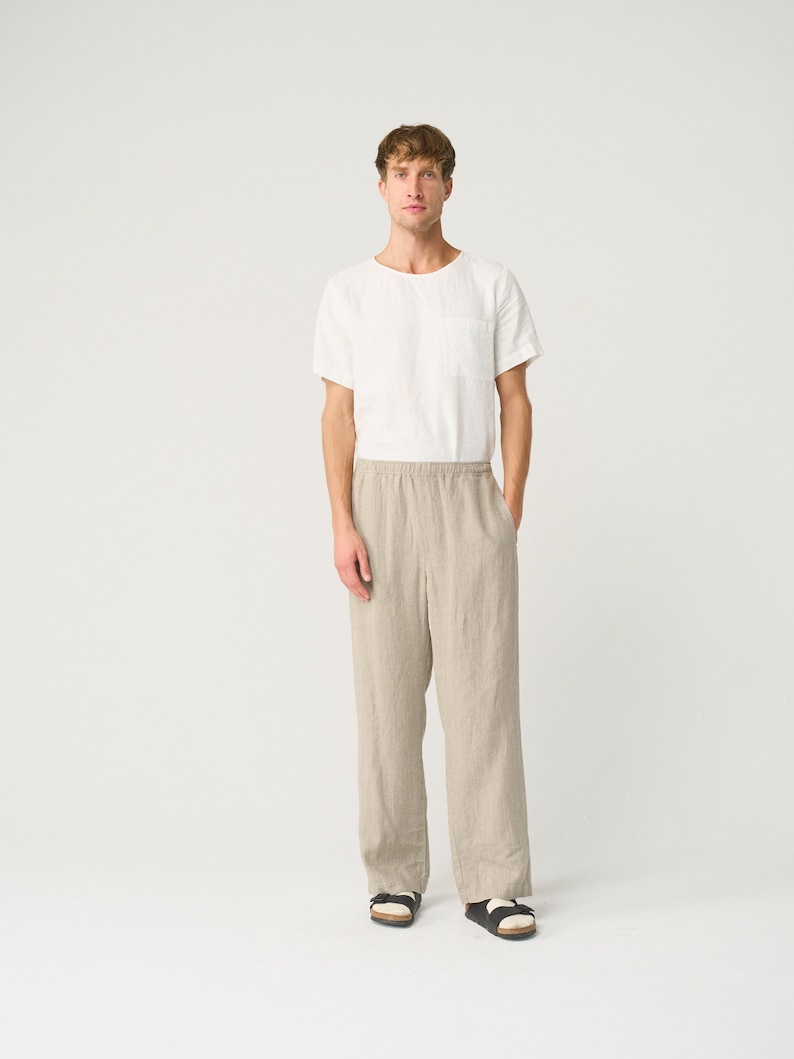 May include: A man is wearing a white linen t-shirt with a pocket on the left side and beige linen pants. He is wearing black sandals.