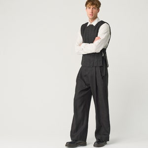 May include: A man wearing a black quilted vest over a white striped shirt, black pants, and black shoes. He is standing with his arms crossed in front of a white background.