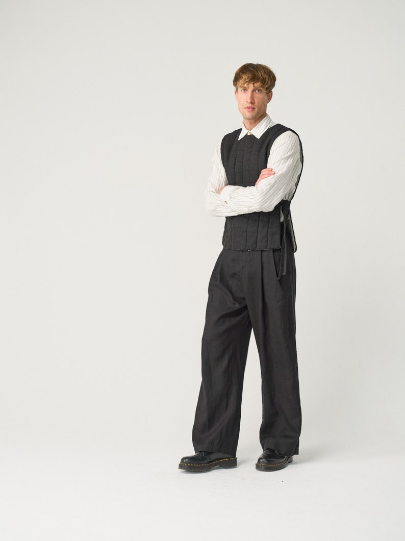 May include: A man wearing a black quilted vest over a white striped shirt, black pants, and black shoes. He is standing with his arms crossed in front of a white background.