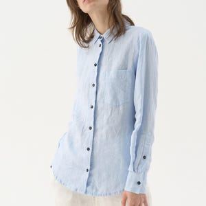 May include: A light blue button-down shirt with a chest pocket and long sleeves. The shirt has black buttons and is made of a lightweight fabric. The shirt is paired with white pants.