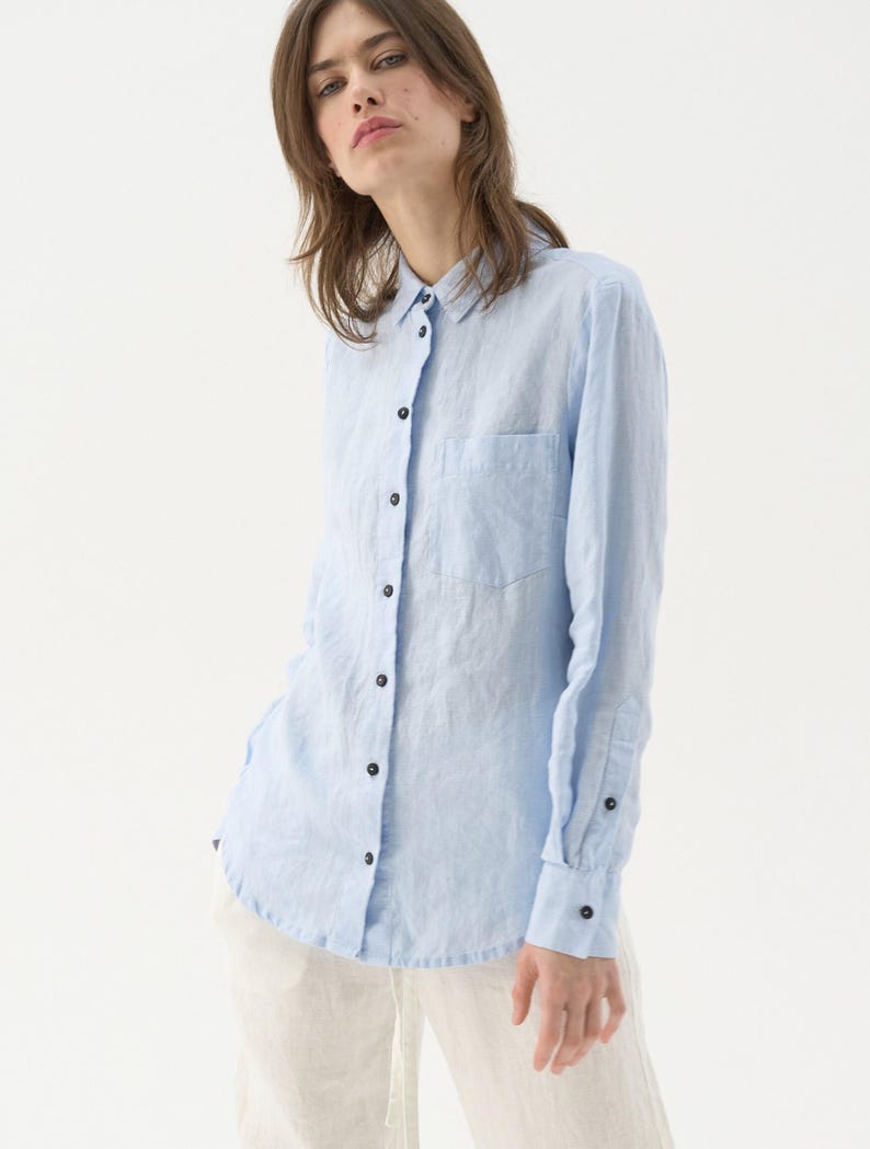 May include: A light blue button-down shirt with a chest pocket and long sleeves. The shirt has black buttons and is made of a lightweight fabric. The shirt is paired with white pants.