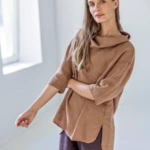 May include: A brown linen top with a cowl neck and three-quarter sleeves. The top has a relaxed fit and side slits. The model is also wearing dark brown pants. The outfit is made of linen fabric.