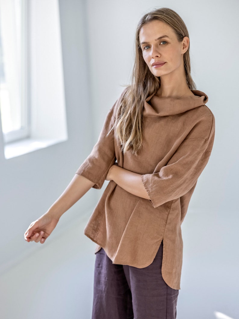 May include: A brown linen top with a cowl neck and three-quarter sleeves. The top has a relaxed fit and side slits. The model is also wearing dark brown pants. The outfit is made of linen fabric.