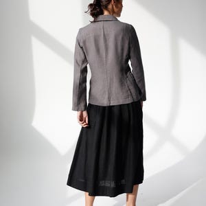 May include: A woman wearing a gray blazer and a black skirt. The blazer has a classic collar and long sleeves. The skirt is a midi length. The woman is wearing brown loafers. The background is white with shadows.