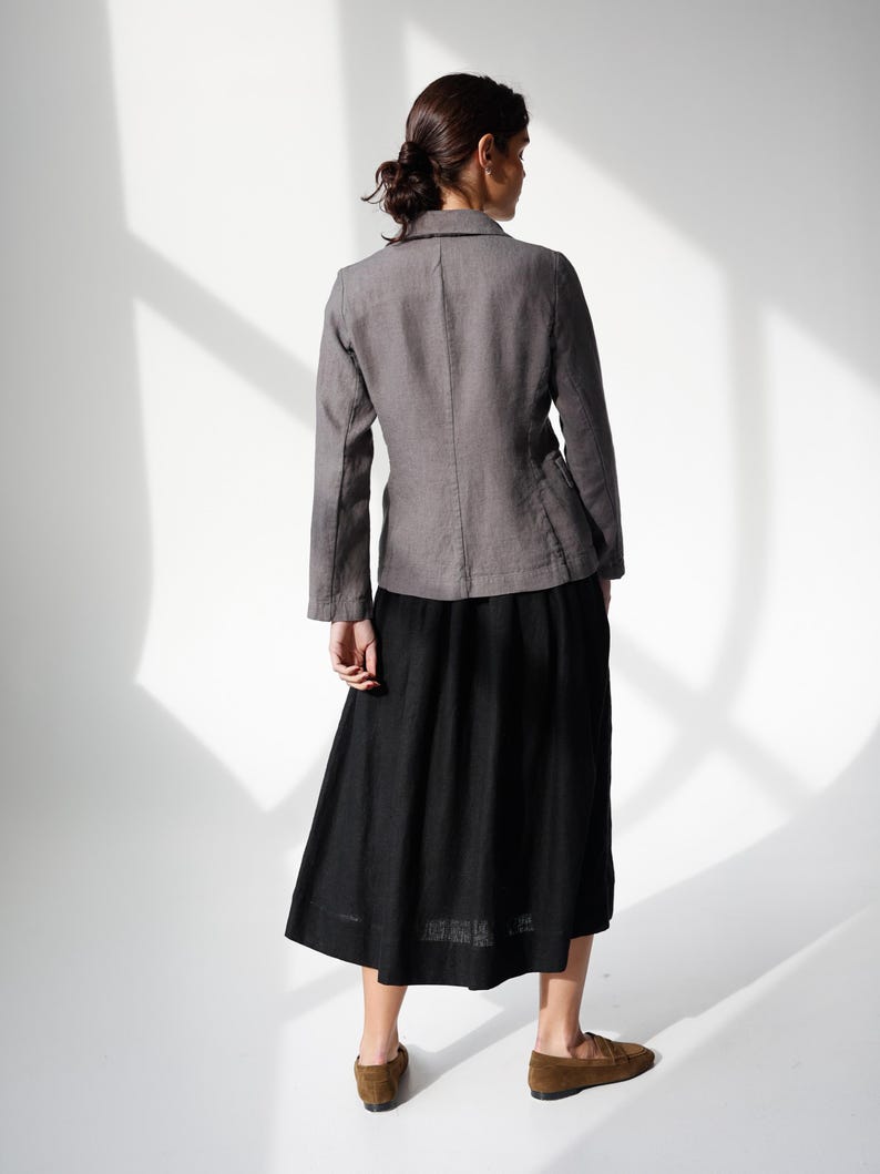 May include: A woman wearing a gray blazer and a black skirt. The blazer has a classic collar and long sleeves. The skirt is a midi length. The woman is wearing brown loafers. The background is white with shadows.