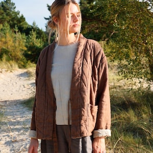 May include: A brown quilted jacket with long sleeves, a collarless open front, and two front pockets. The jacket features visible stitching patterns across the fabric and is worn over a light beige shirt and dark gray trousers.
