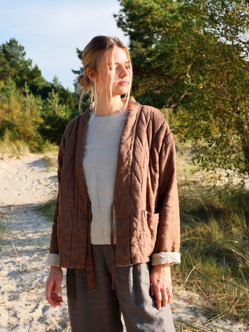 May include: A brown quilted jacket with long sleeves, a collarless open front, and two front pockets. The jacket features visible stitching patterns across the fabric and is worn over a light beige shirt and dark gray trousers.