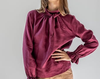 May include: A burgundy long-sleeved blouse with a tie-neck detail and ruffled cuffs. The blouse is paired with tan-colored pants. The outfit is made of a soft, flowing fabric, suggesting a comfortable and stylish look.