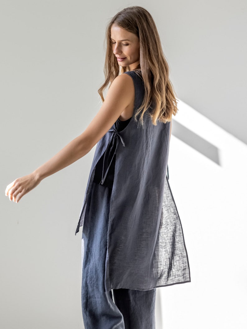 Puede incluir: Una blusa larga de lino de color gris antracita sin mangas con un lazo en la cintura. La blusa tiene una espalda larga y fluida con una abertura a cada lado.