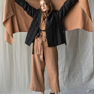 May include: A woman wearing a black quilted jacket, a tan jumpsuit, and black shoes. She is holding up a large, tan-colored fabric. The outfit is a neutral color palette, with a focus on natural materials and textures.