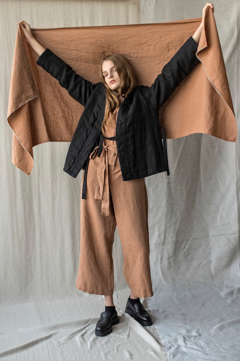 May include: A woman wearing a black quilted jacket, a tan jumpsuit, and black shoes. She is holding up a large, tan-colored fabric. The outfit is a neutral color palette, with a focus on natural materials and textures.