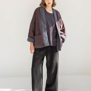 May include: A person wearing a quilted, burgundy jacket with blue trim and pockets, paired with a dark blue top and black wide-leg trousers. The jacket has a relaxed fit, and the outfit is set against a white background.