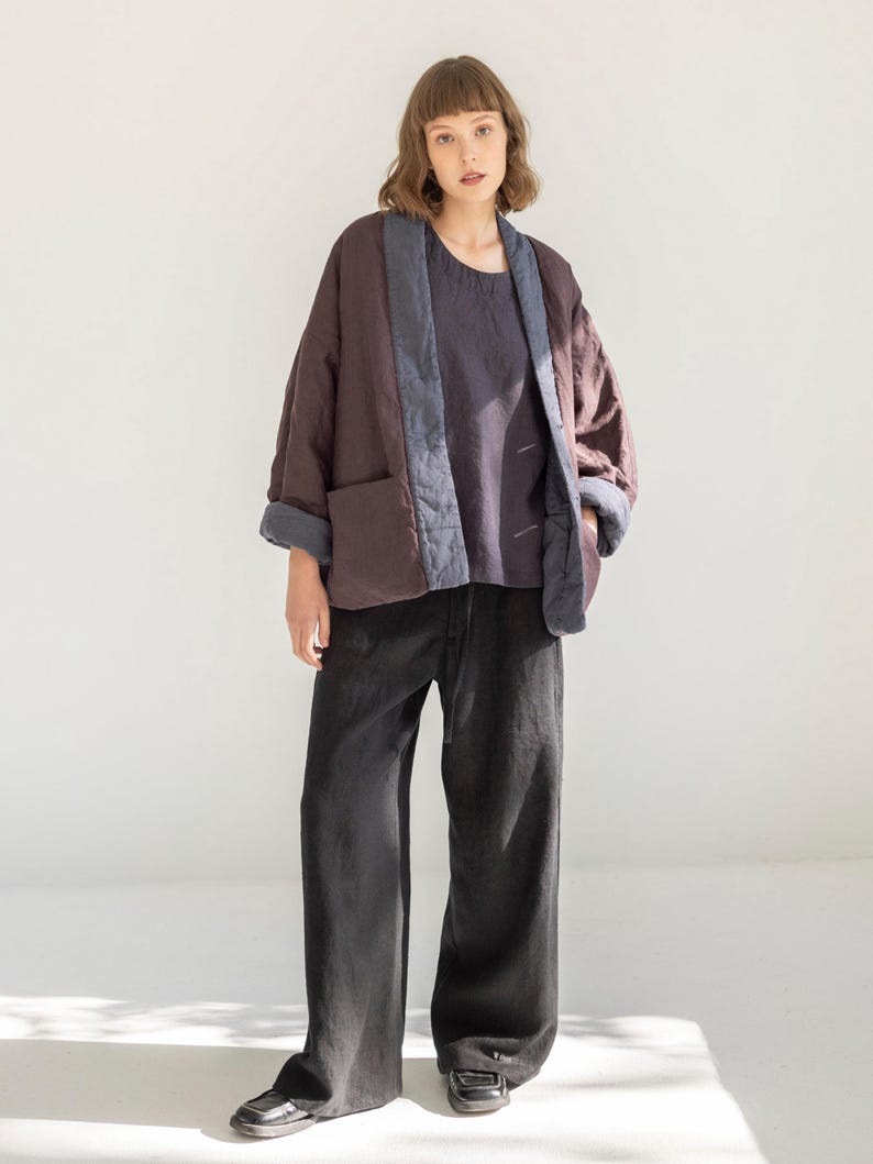 May include: A person wearing a quilted, burgundy jacket with blue trim and pockets, paired with a dark blue top and black wide-leg trousers. The jacket has a relaxed fit, and the outfit is set against a white background.