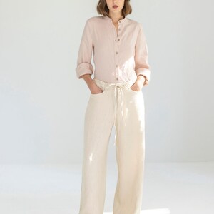 May include: A light pink linen shirt with a mandarin collar and button-down front is paired with cream-colored wide-leg pants. The pants have a drawstring waist and pockets. The outfit is completed with beige sneakers.