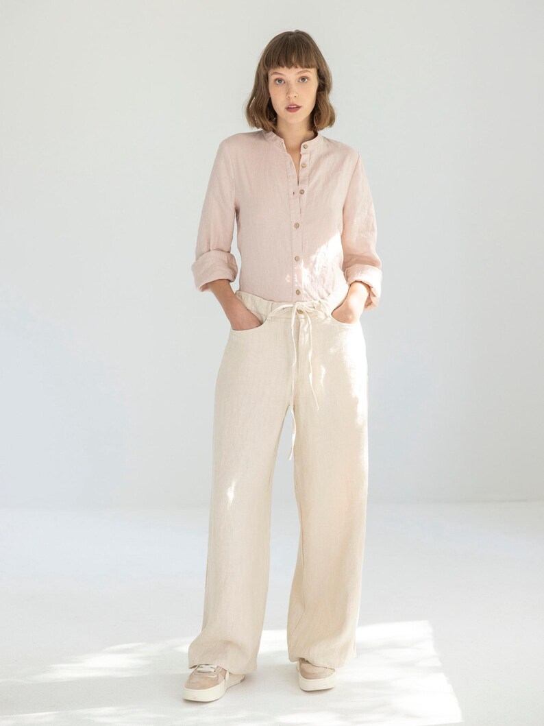 May include: A light pink linen shirt with a mandarin collar and button-down front is paired with cream-colored wide-leg pants. The pants have a drawstring waist and pockets. The outfit is completed with beige sneakers.