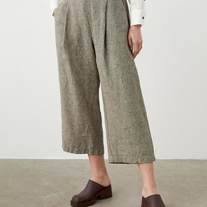 K&ouml;nnte beinhalten: Eine Hose mit weitem Bein und kurzem Schnitt aus strukturiertem, neutralem Stoff. Die Hose hat Falten an der Taille und Taschen. Das Model tr&auml;gt braune Clogs. Ein wei&szlig;es Hemd mit Kn&ouml;pfen ist teilweise sichtbar.